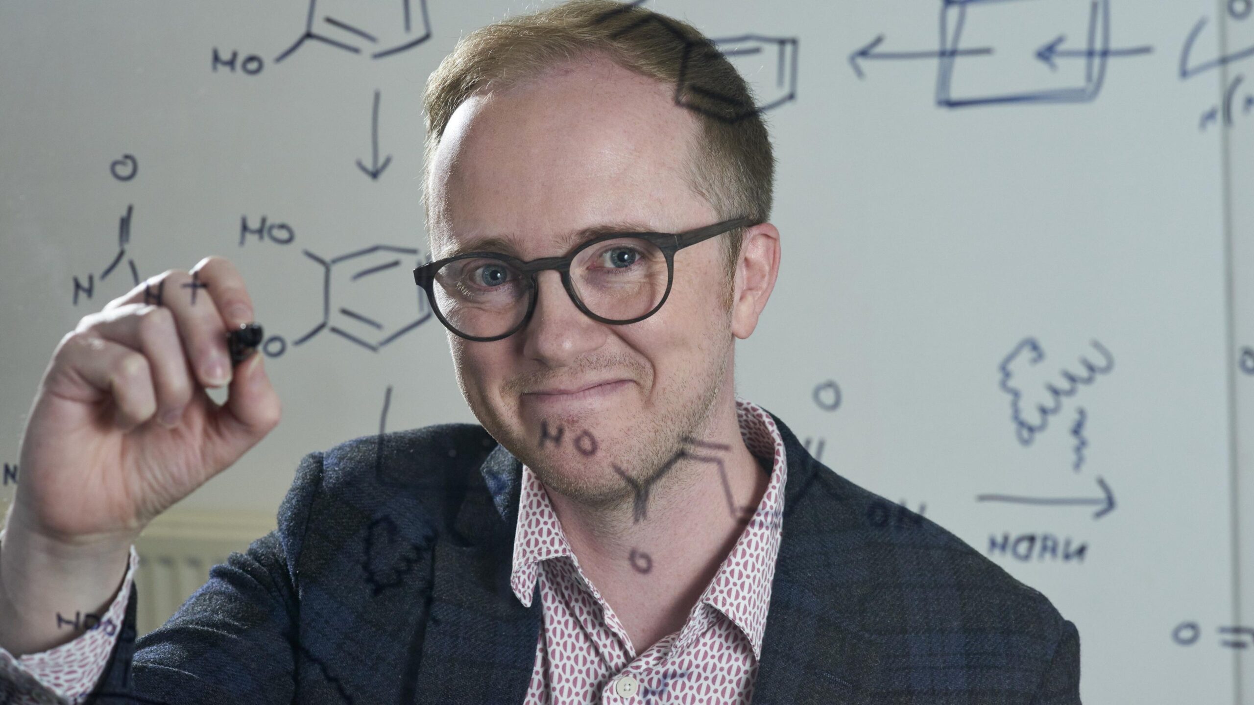 Smiling Professor Stephen Wallace writes chemical formulae on a see-through display board.