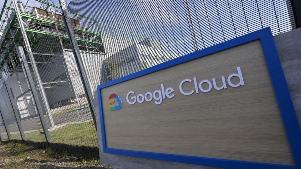 A Google Cloud sign sits in front of a wire fence. Behind is a data centre.
