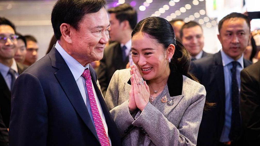 A smiling Paetongtarn Shinawatra turns to her father and former Thai Prime Minister Thaksin Shinawatra with her hands folded in a gesture of respect. They are at a public event surrounded by other officials. Thaksin is wearing a navy blue suit with a pink tie and looking ahead, half-smiling. Paetongtarn is wearing a grey suit. 