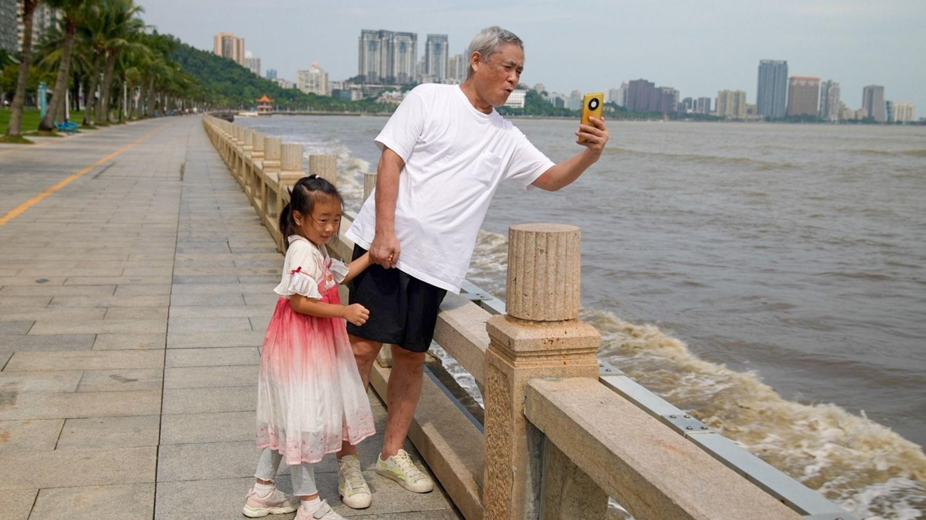 A man in a t-shirt and shorts takes a photo of the ocean while a little girl in a dress holds his hand.