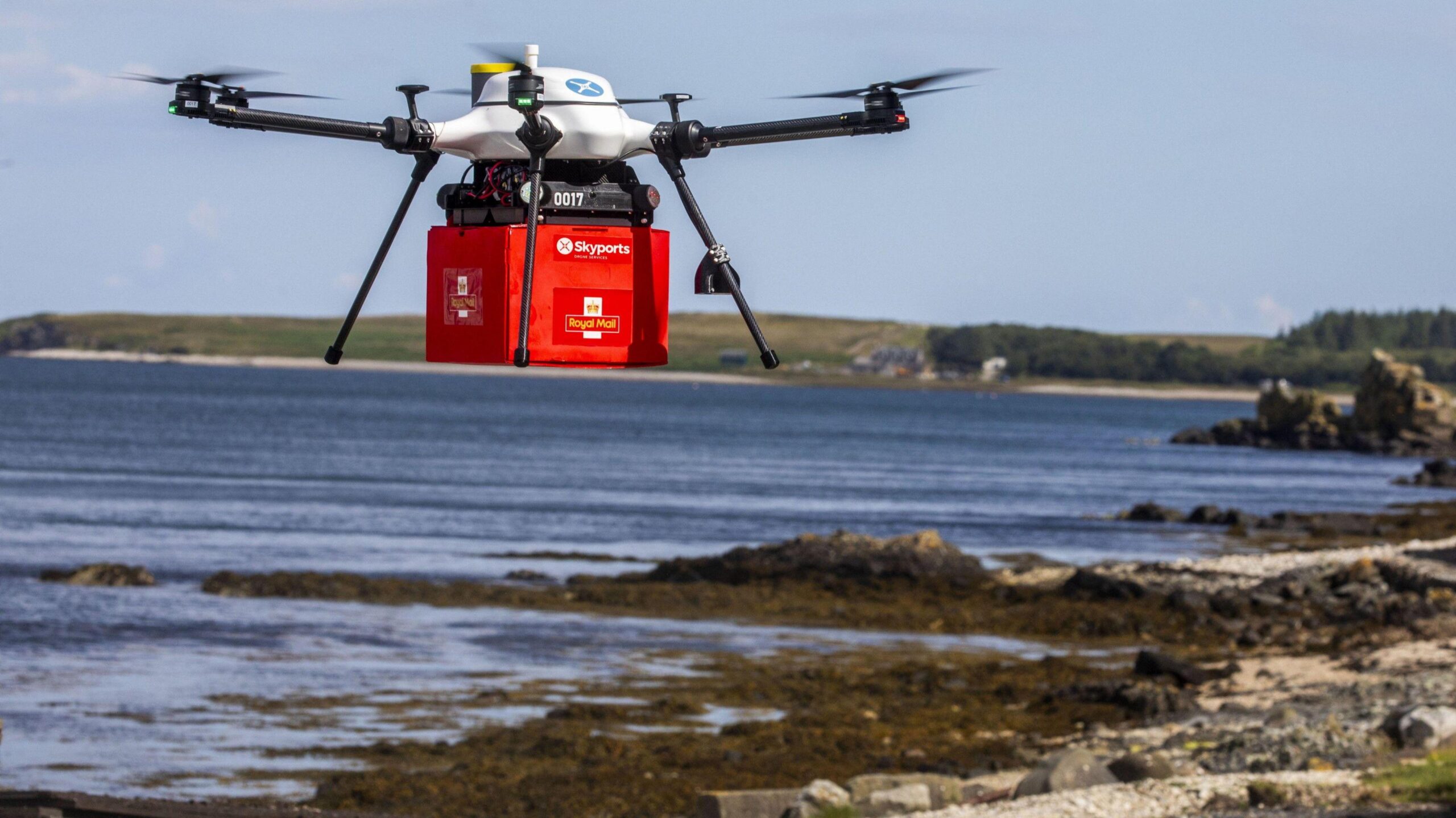 A white Skyports drones carries a red box with the Royal Mail logo, over a beach with the sea in the background.