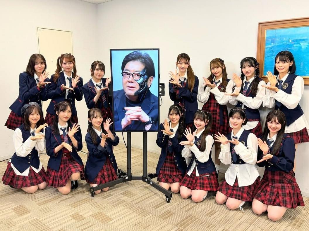 Members of AKB48 pose next to a screen showing an "AI" image of their composer Yasushi Akimoto