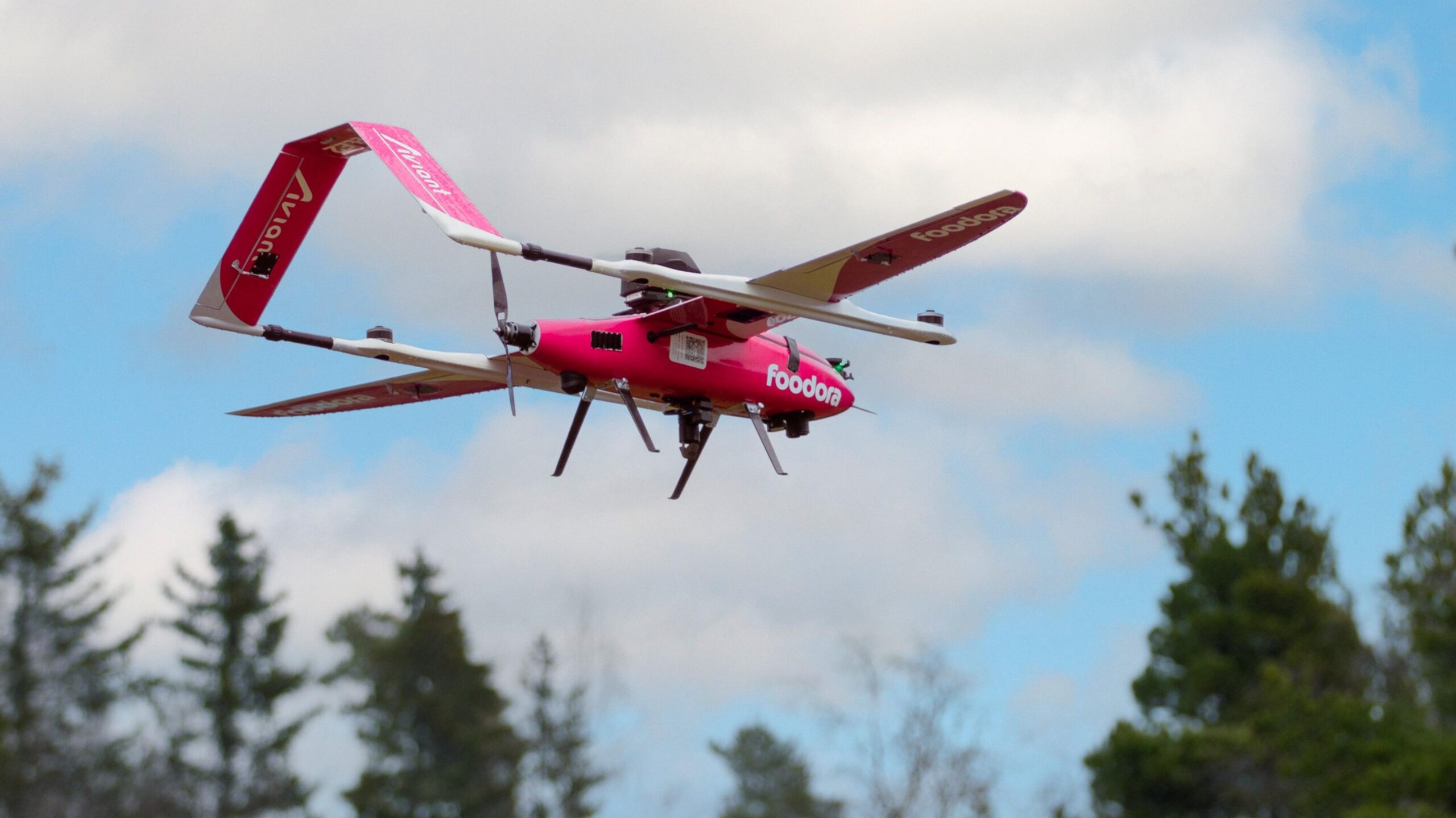 The pink Foodora drone flying against the sky with treetops in the distance.