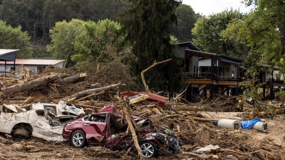 Fallen trees and debris on top of a crumpled red and white car. There is a house in the background largely intact.