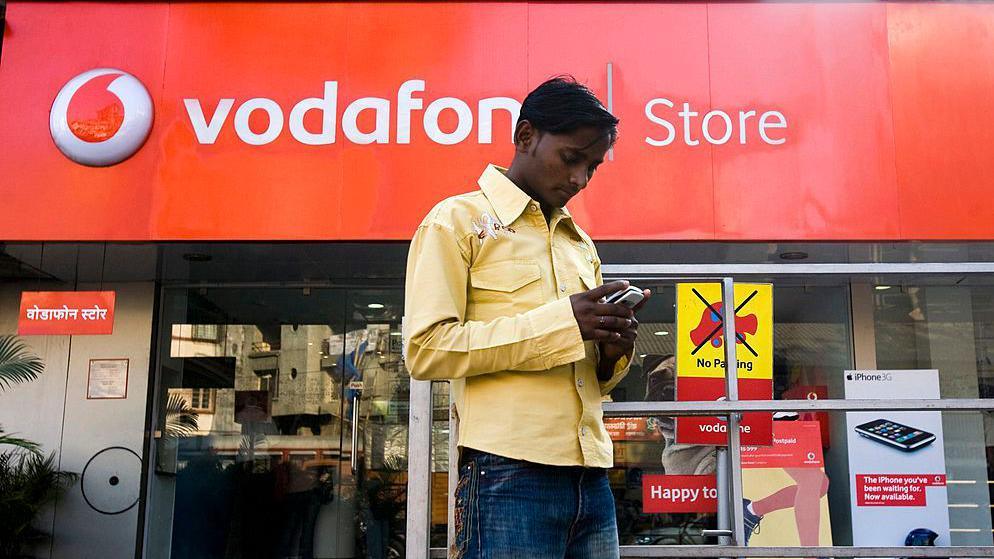 A pedestrian wearing a yellow shirt and blue jeans uses his mobile phone outside a Vodafone store in Mumbai