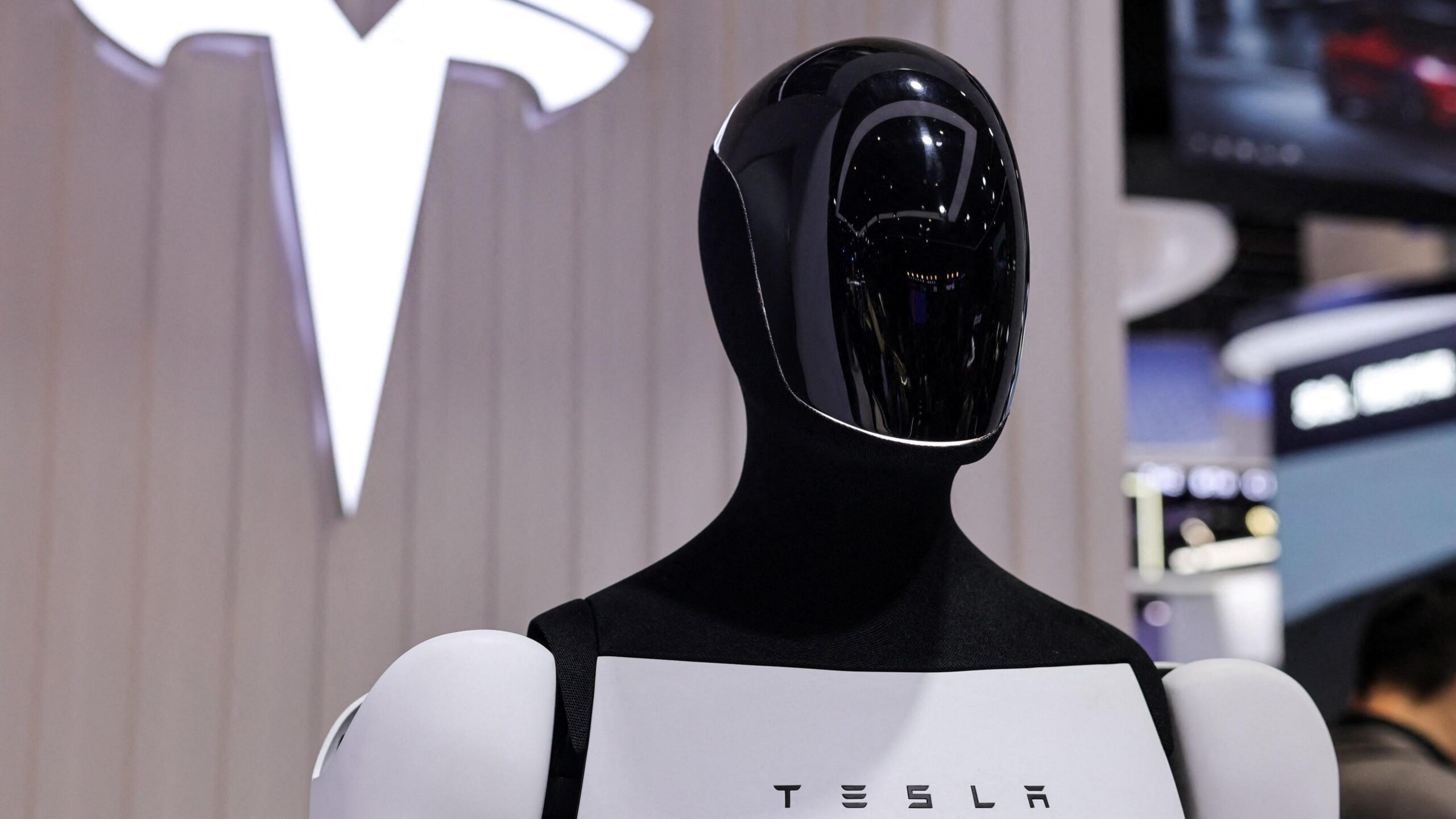 Tesla's Optimus humanoid robot stands in front of a sign displaying the company's logo.