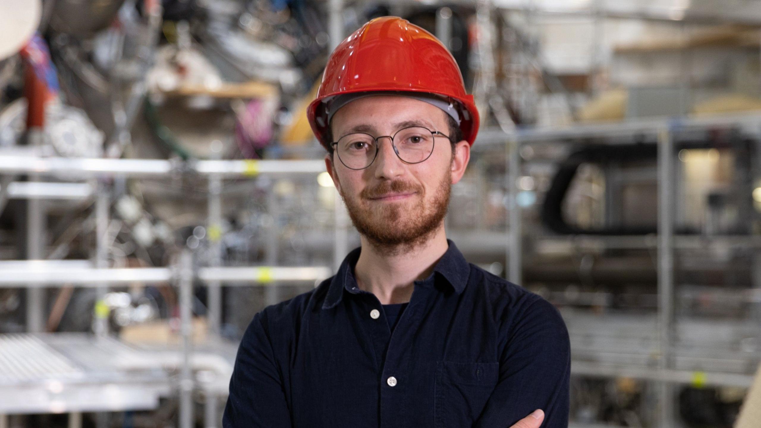 Wearing a red safety helmet and round glasses, Francesco Sciortino stands in front of fusion equipment.
