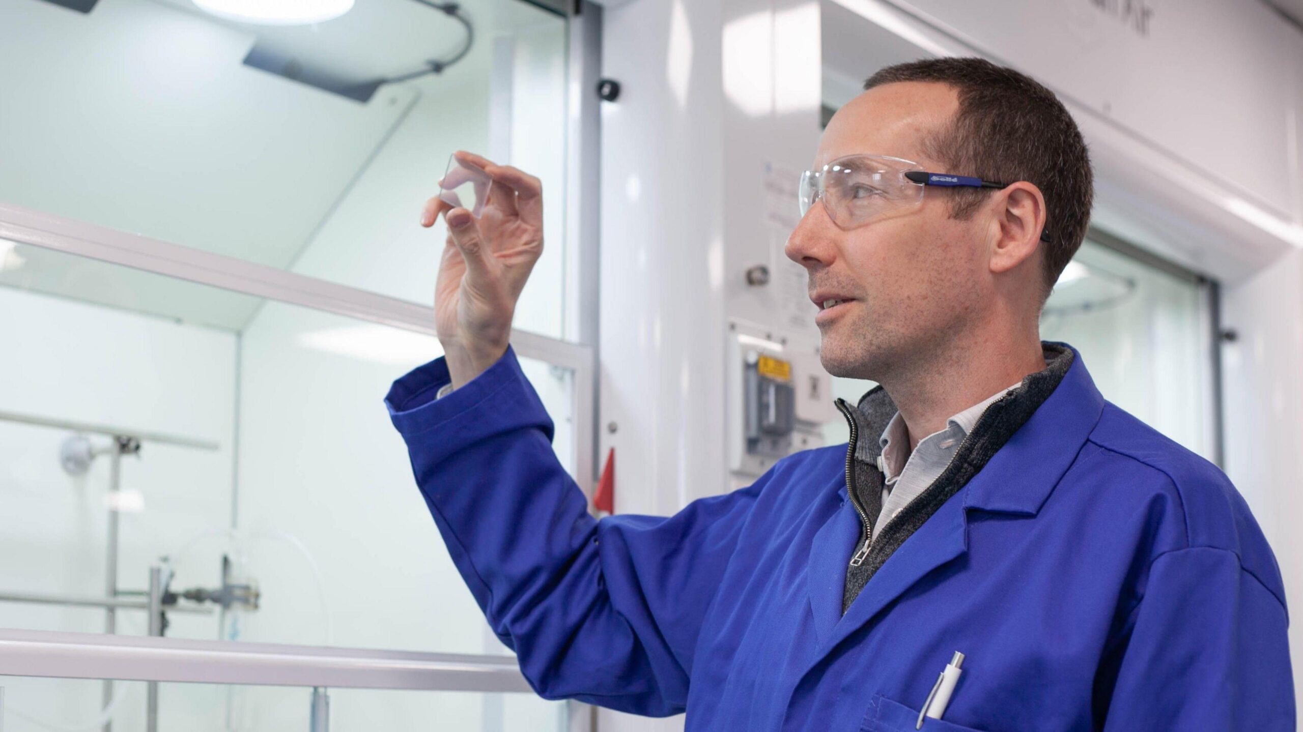 John Griffin, wearing a blue lab coat and safety specs, looks at a glass slide.