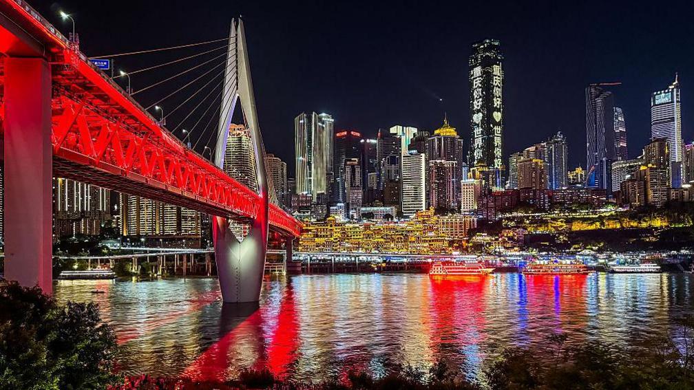 Tourists gather along the riverside promenade near the illuminated Qiansimen Bridge, with Hongya Cave and the modern skyscraper skyline of Chongqing's central business district in front of them,.
