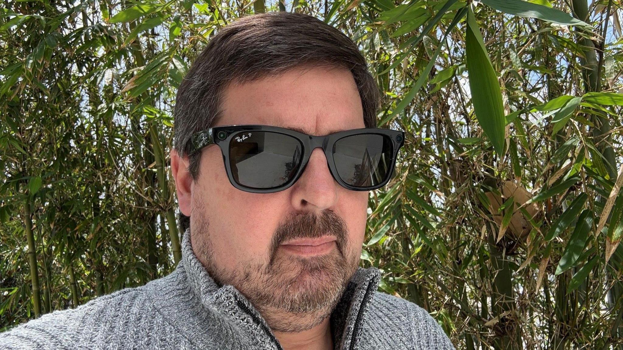 A man standing outside in a grey sweater and wearing Meta's Ray-Bans with a wall of green bamboo behind him. 